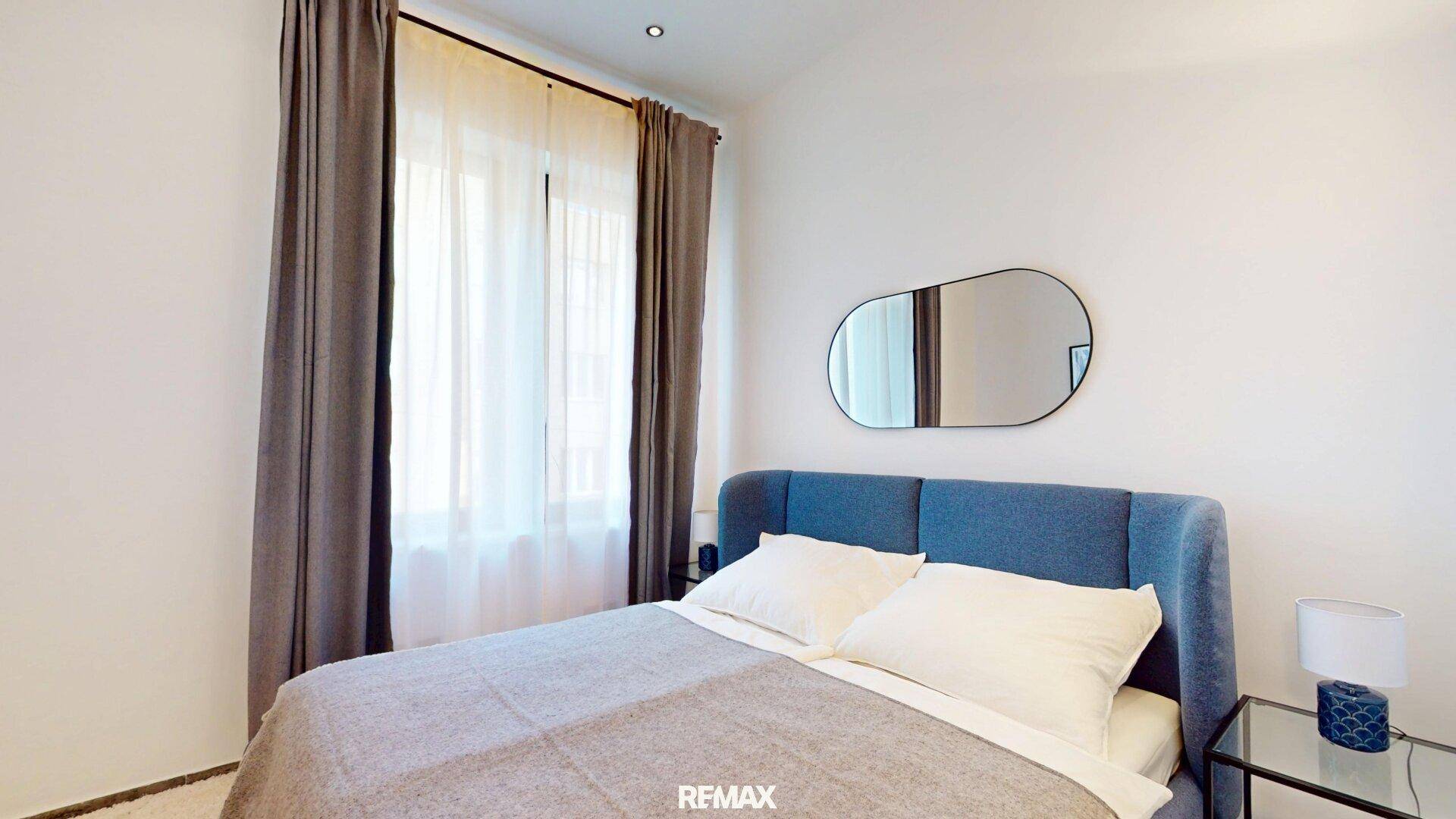 Gemütliches Schlafzimmer mit blauem Polsterbett, großem Fenster und modernem Spiegel.