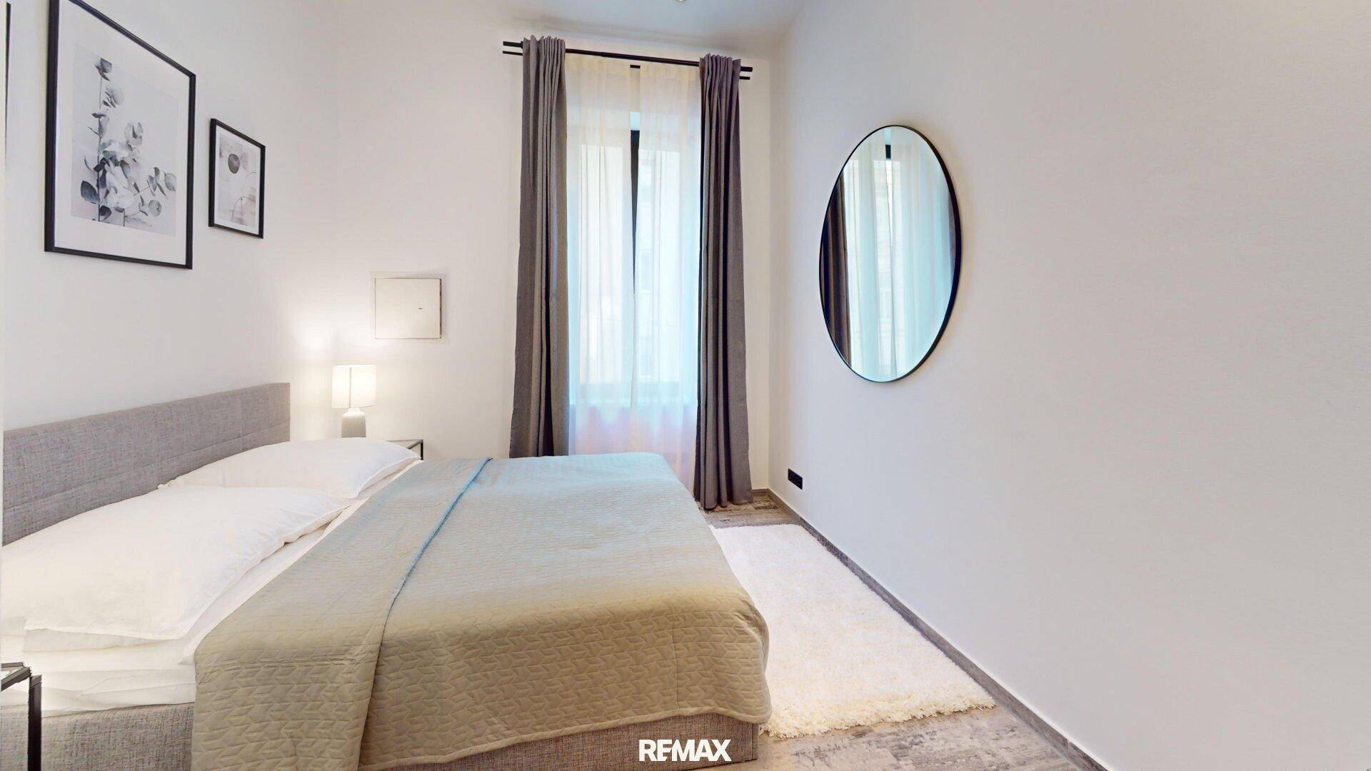 Helles Schlafzimmer mit grauem Bett, großem Fenster und rundem Wandspiegel.
