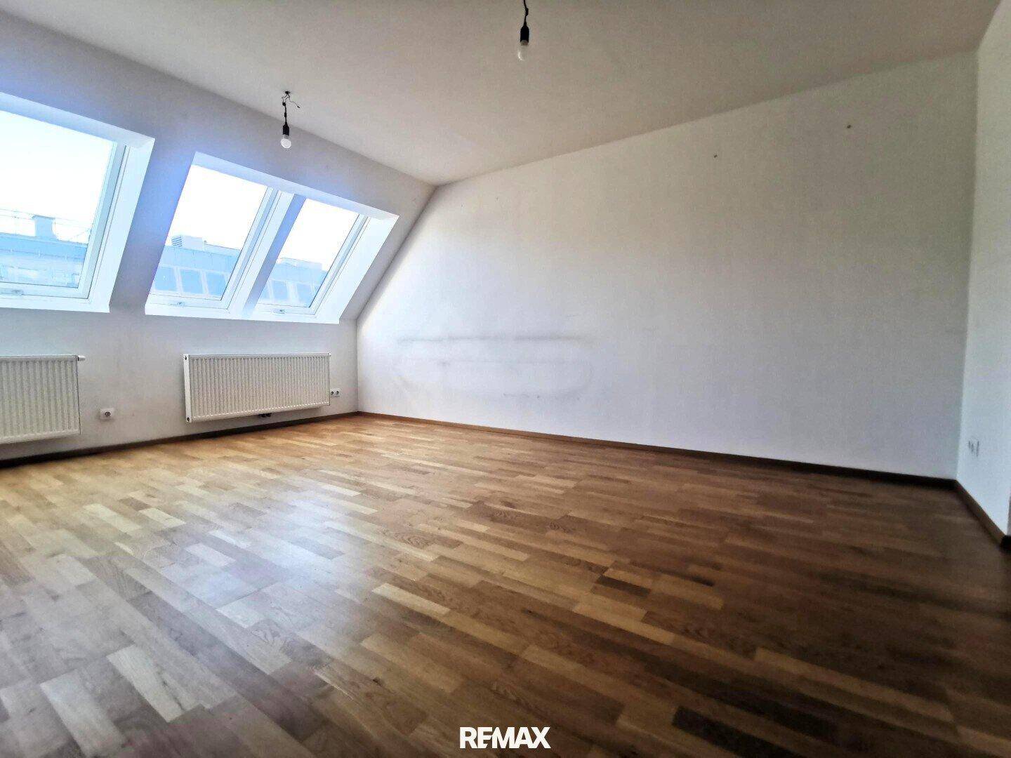 Helles Dachzimmer mit Holzboden, schrägen Wänden und zwei großen Dachfenstern für viel Tageslicht.