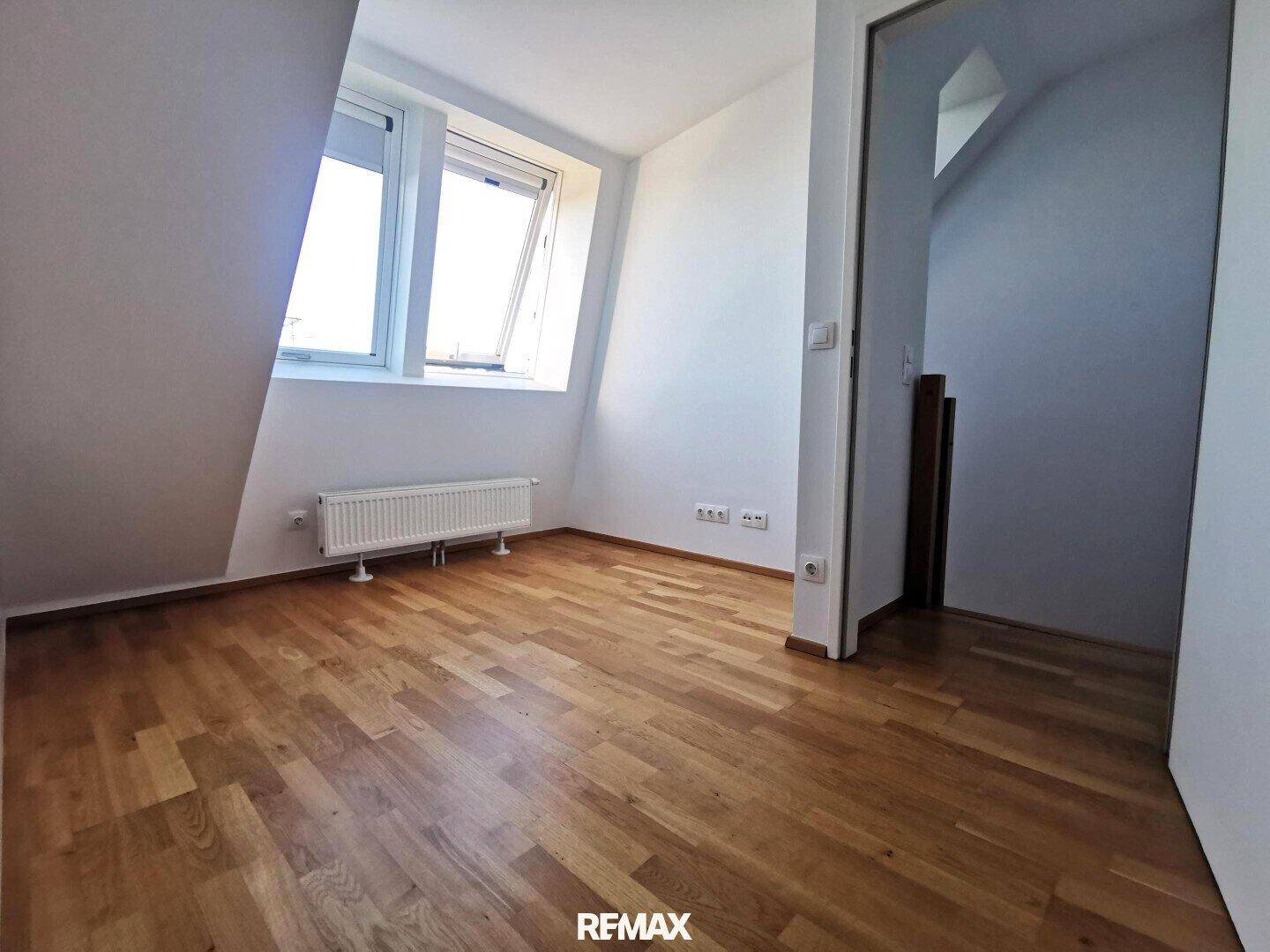 Helles Zimmer mit Parkettboden, weißen Wänden, Fenster und Heizkörper unter einer Dachschräge.