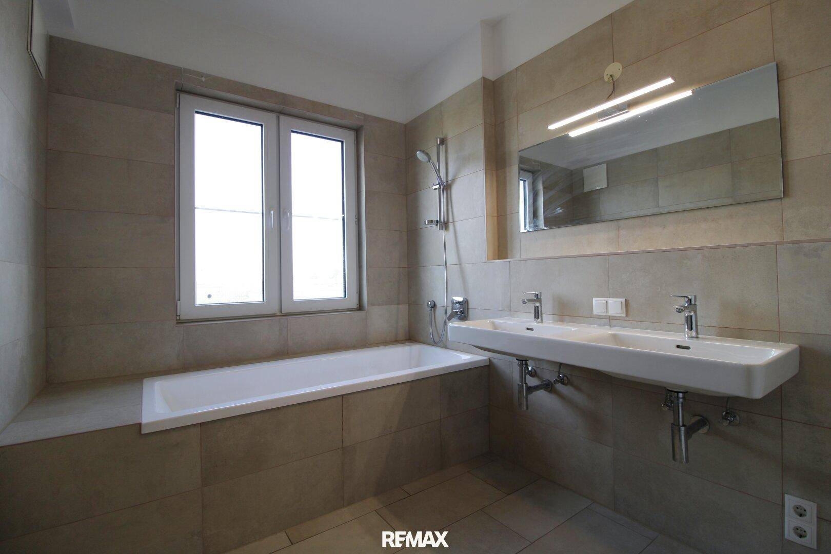 Modernes Badezimmer mit Badewanne, Doppelwaschbecken, Fenster und beigefarbenen Fliesen.