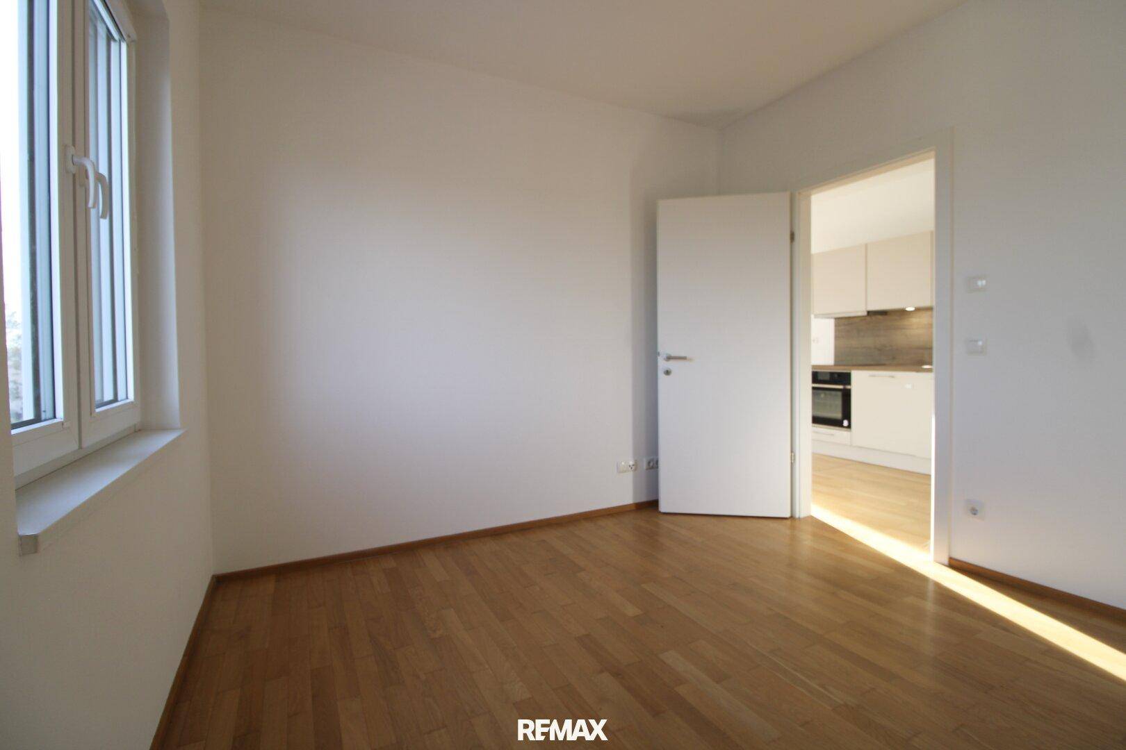 Geräumiges Zimmer mit Parkettboden und Fenster, mit Blick auf die angrenzende Küche.