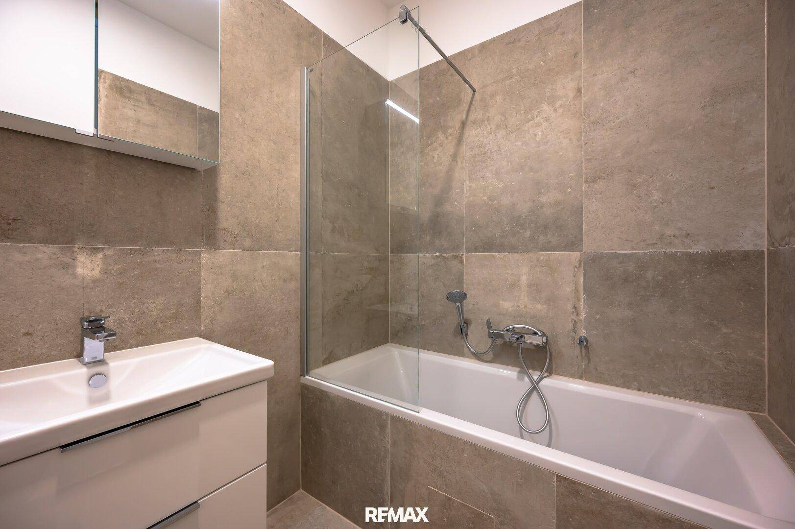 Modernes Badezimmer mit Badewanne, Duschwand, Waschtisch und großformatigen grauen Fliesen.