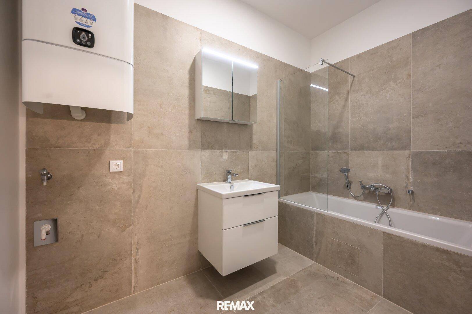 Helles Badezimmer mit Badewanne, modernem Waschtisch, Warmwasserbereiter und grauen Wandfliesen.