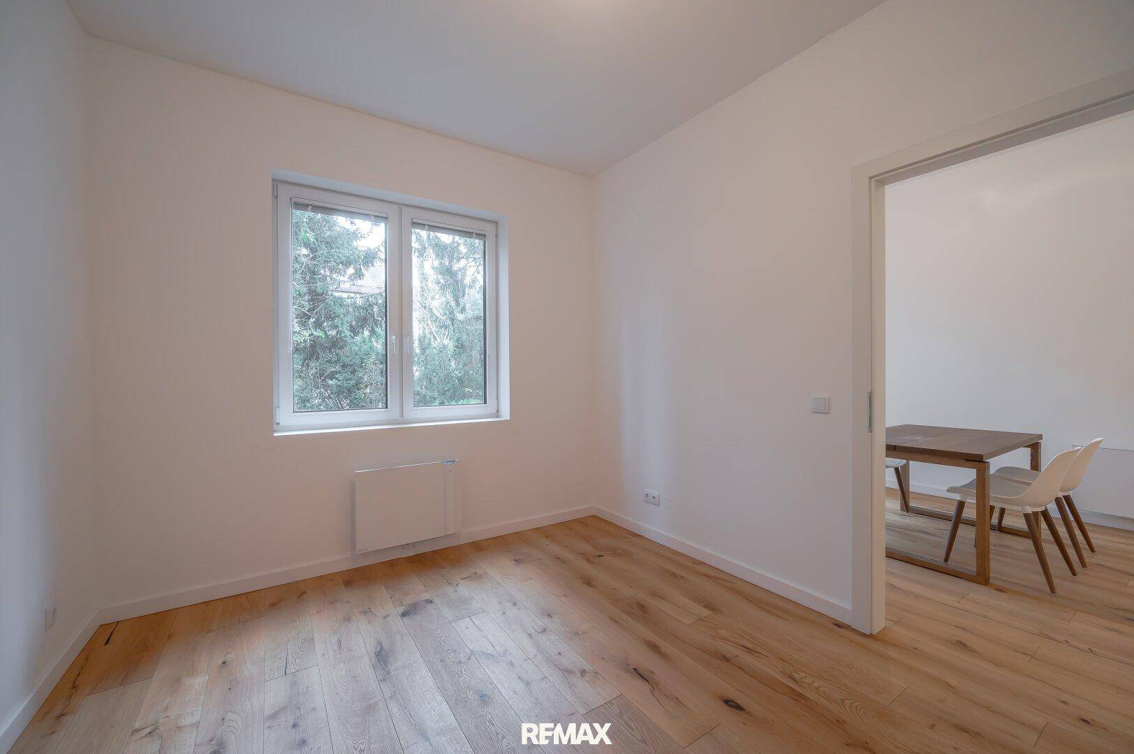 Helles Zimmer mit Holzboden, weißen Wänden und großem Fenster mit Blick ins Grüne.