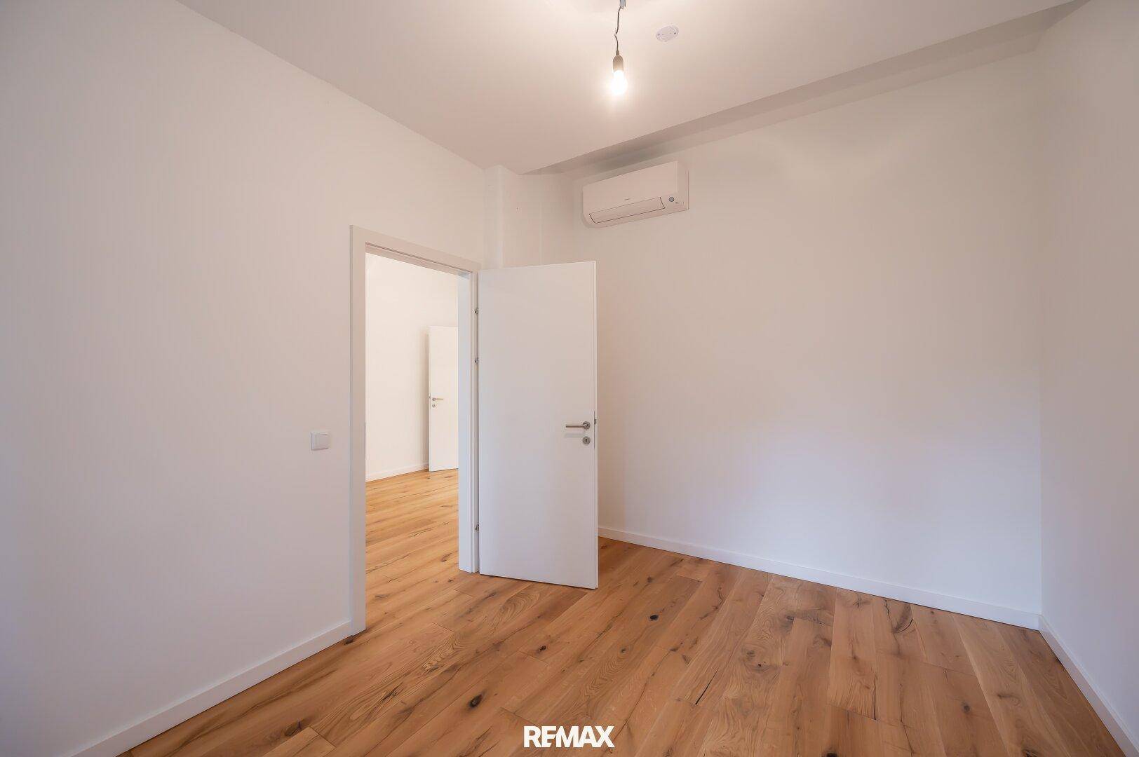 Geräumiges Zimmer mit Holzboden, weißen Wänden und Klimaanlage, mit Zugang zu einem weiteren Raum.