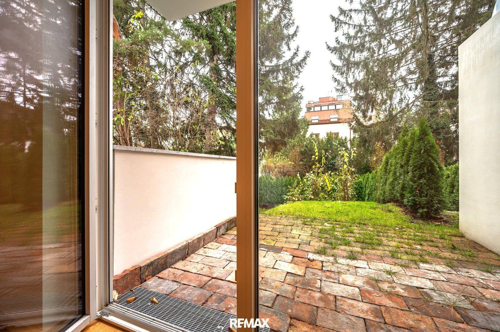 Blick durch die große Glastür auf die gepflasterte Terrasse und den grünen Gartenbereich.