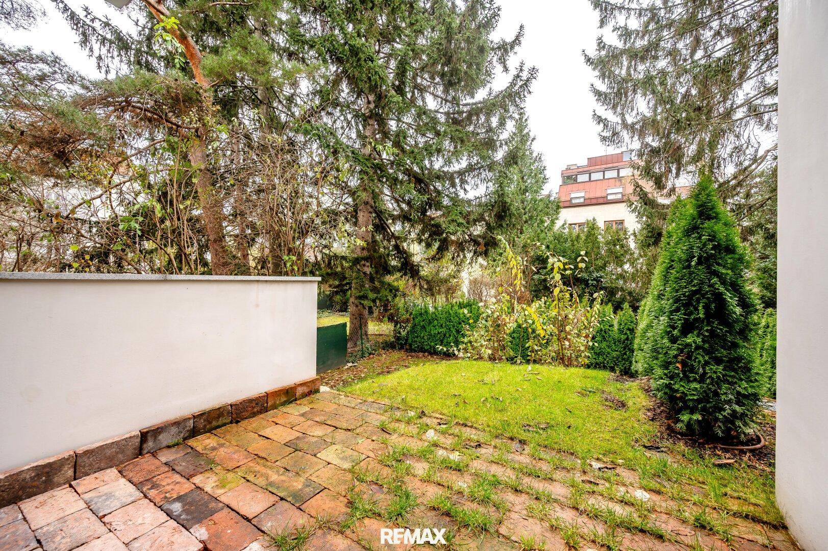 Kleiner gepflasterter Terrassenbereich mit Rasenfläche und umgebenden Bäumen im Garten.