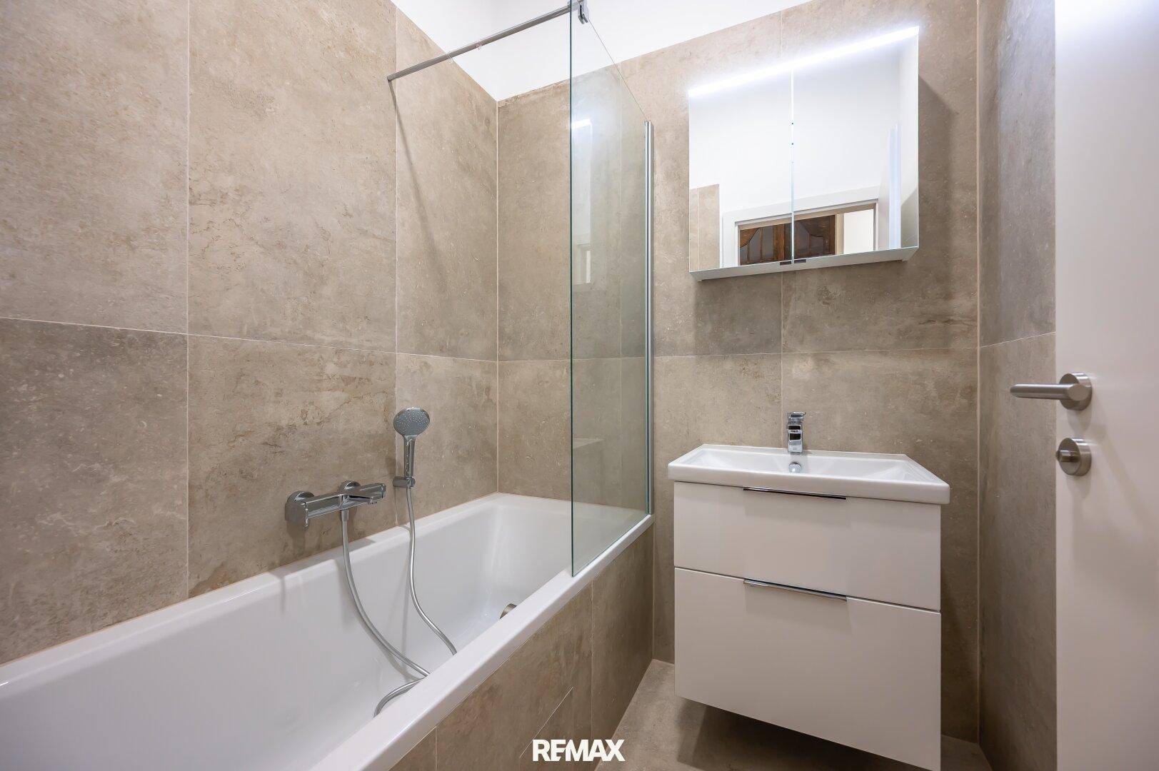 Modernes Badezimmer mit Badewanne, Duschwand, Waschtisch und beleuchtetem Spiegelschrank.