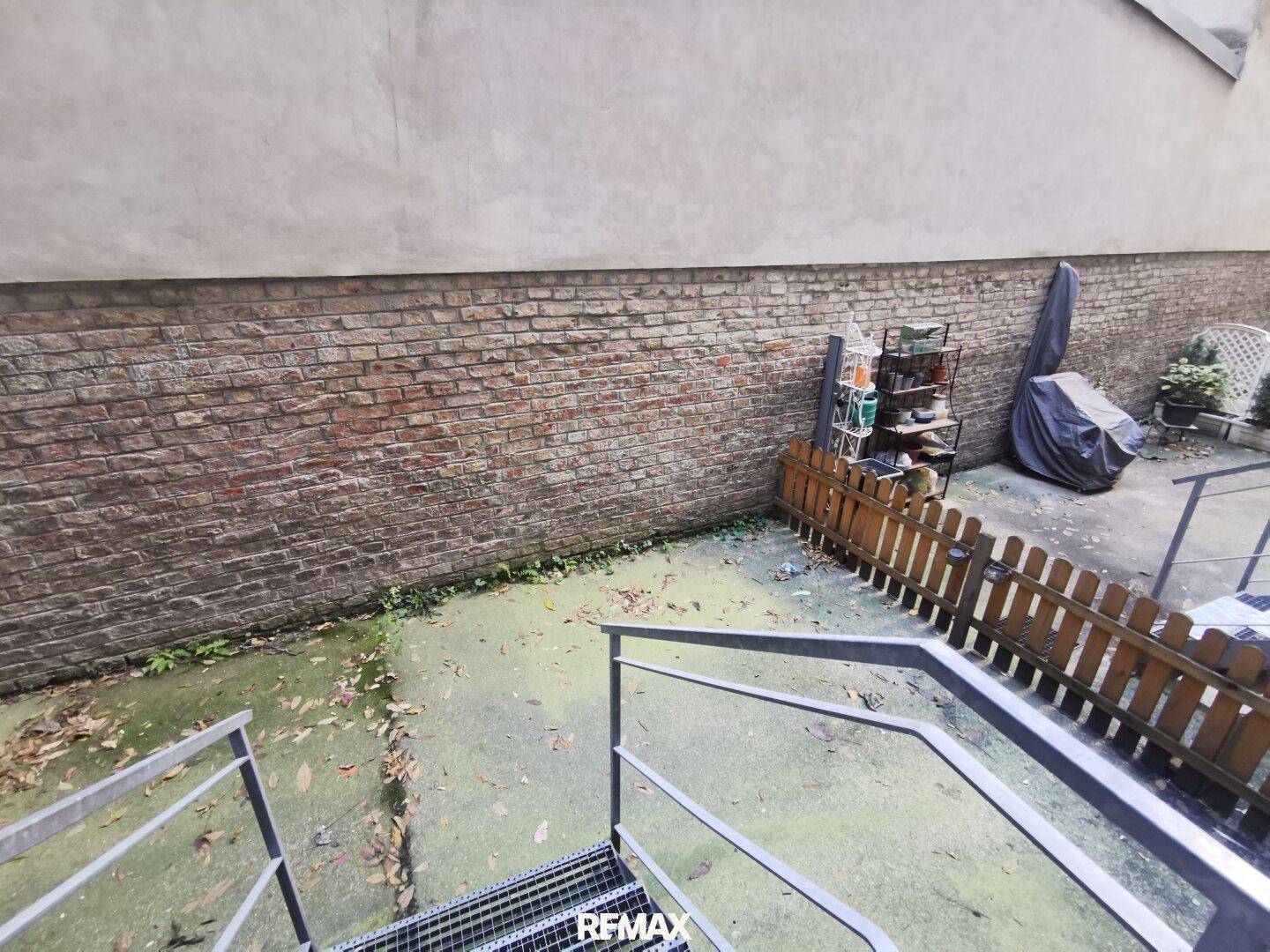Hinterhofbereich mit Ziegelwand, Metalltreppe und einem kleinen, eingezäunten Gartenabschnitt.