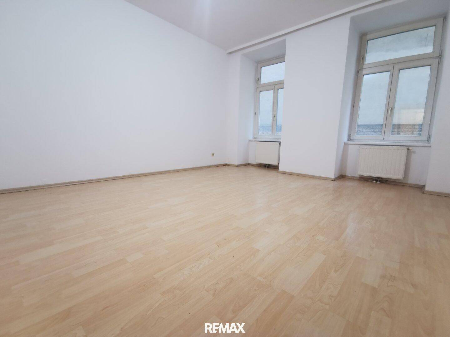 Heller Raum mit Laminatboden und zwei großen Fenstern, die viel Tageslicht hereinlassen.