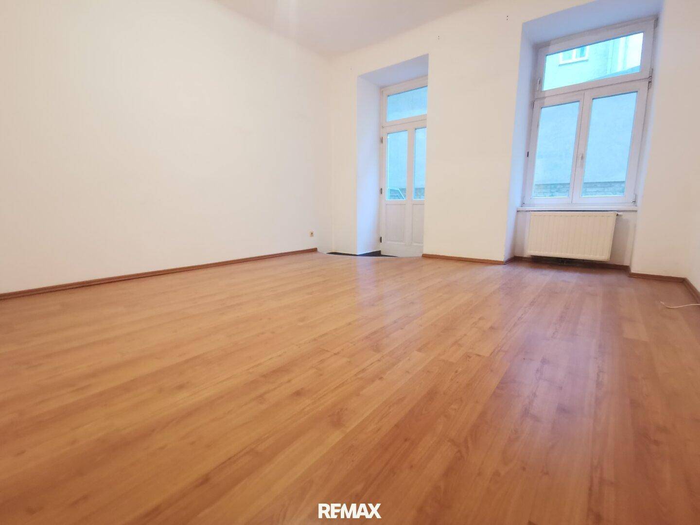 Geräumiger, heller Raum mit Laminatboden, hohen Fenstern und einer Tür, die nach draußen führt.