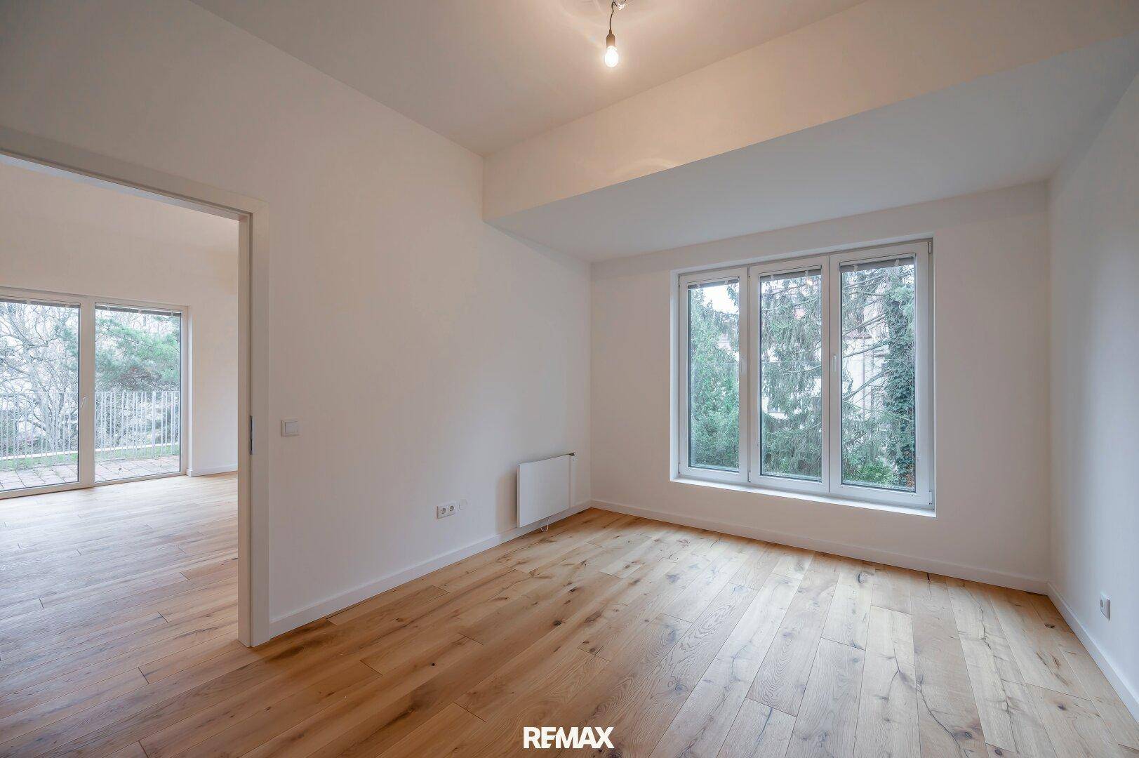 Helles, leeres Zimmer mit Holzboden und großen Fenstern mit Blick ins Grüne.