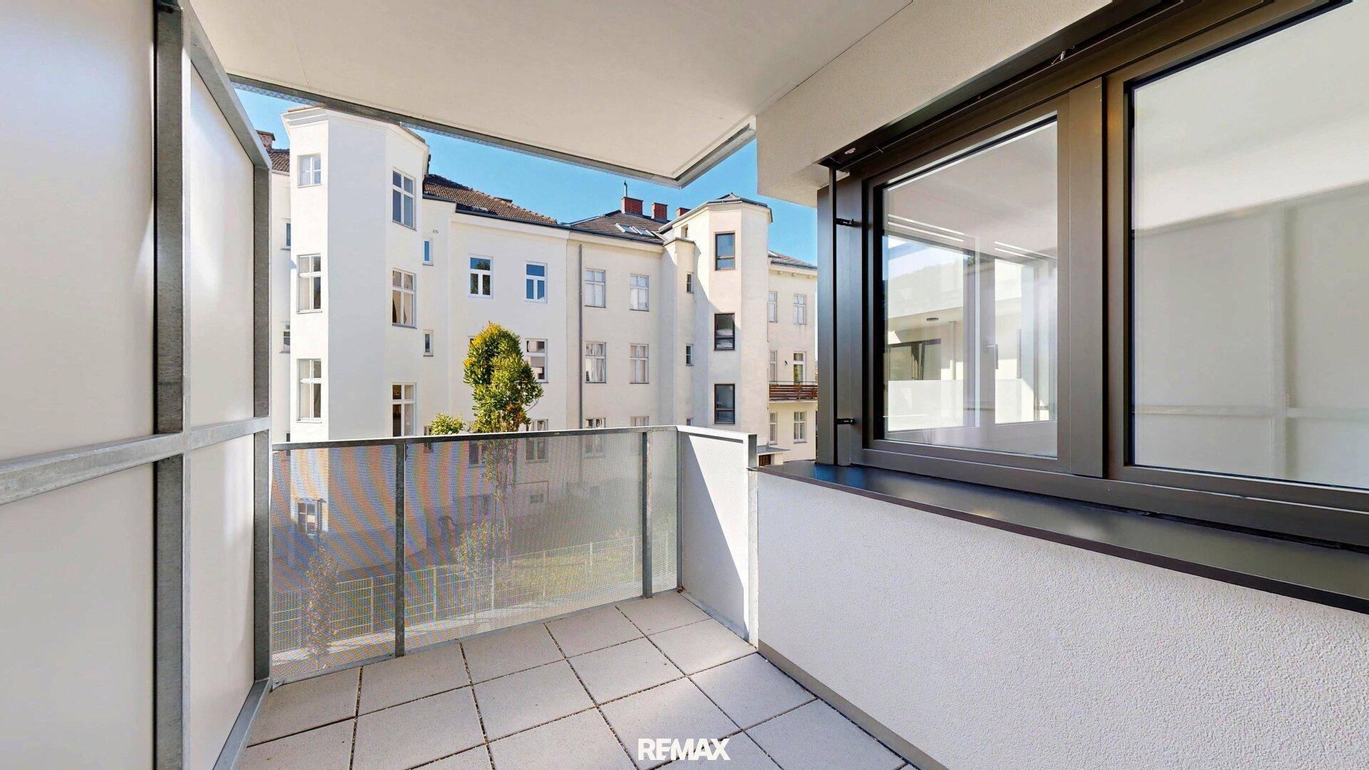 Heller Balkon mit Fliesenboden und Glasgeländer, umgeben von Stadtgebäuden.