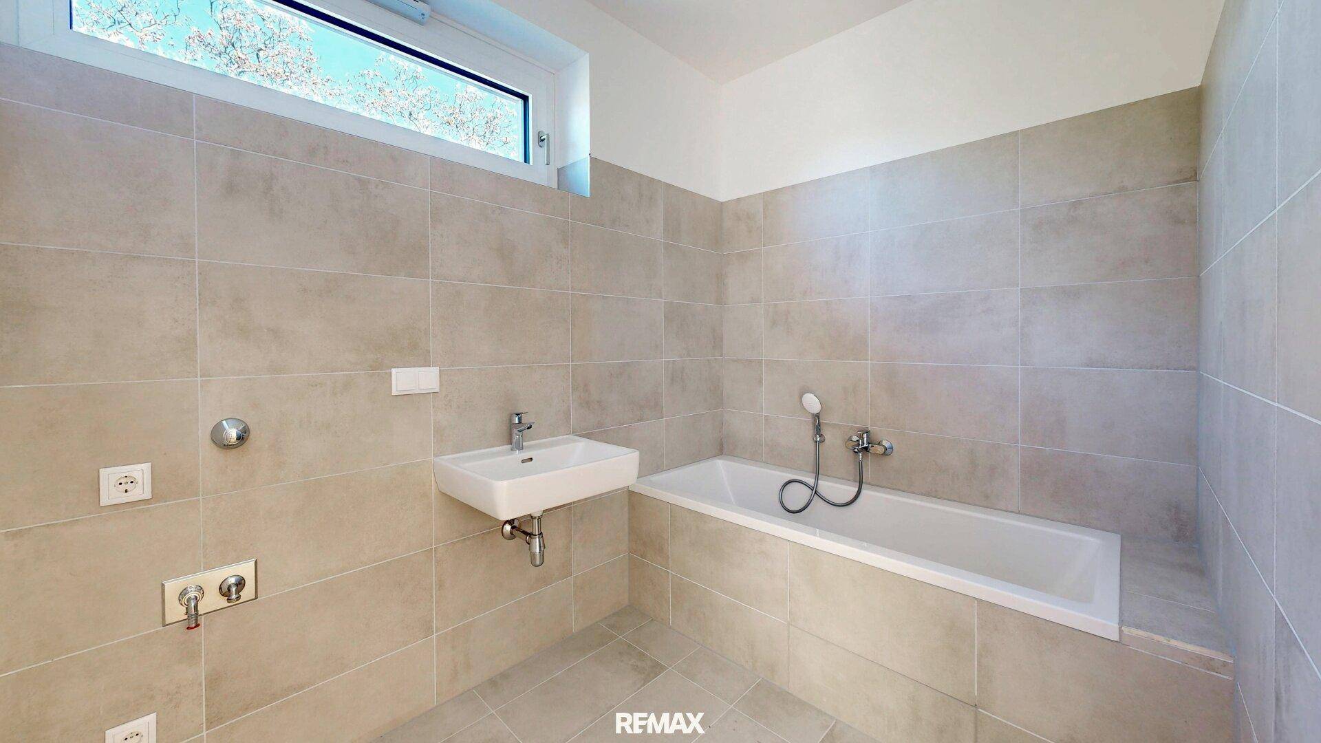 Modernes Badezimmer mit hellen Fliesen, einem Waschbecken und einer Badewanne unter einem Fenster.