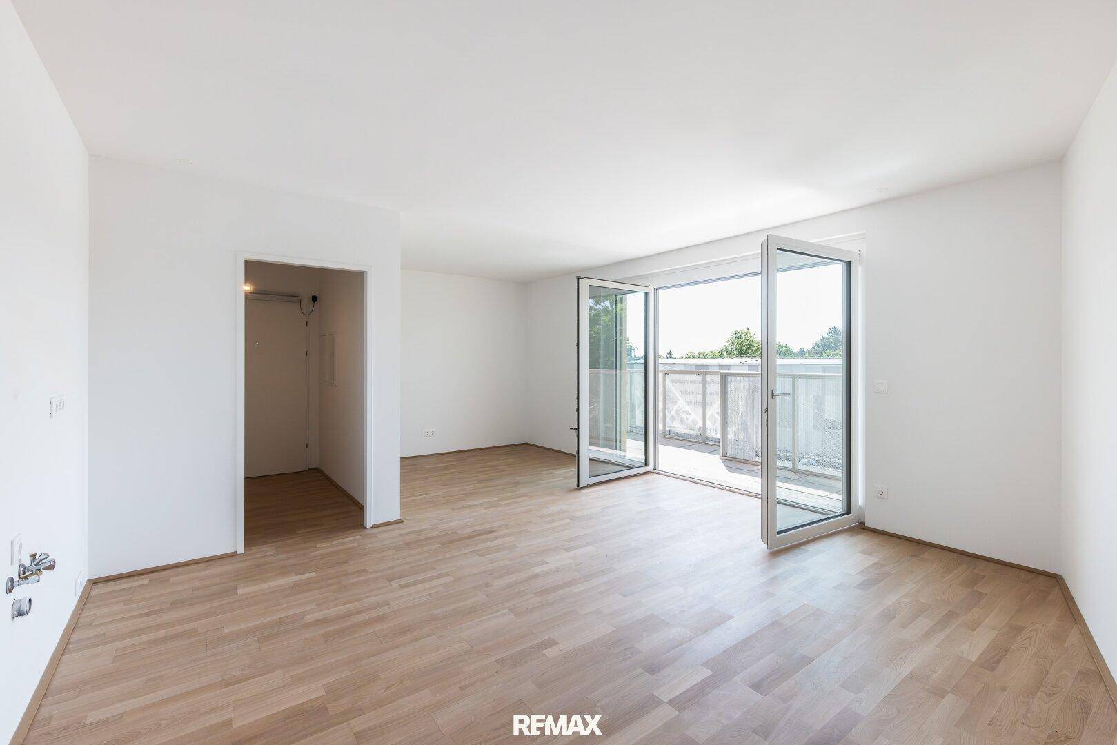 Geräumiges, leeres Wohnzimmer mit hellem Holzboden und großen Fenstertüren zum Balkon.