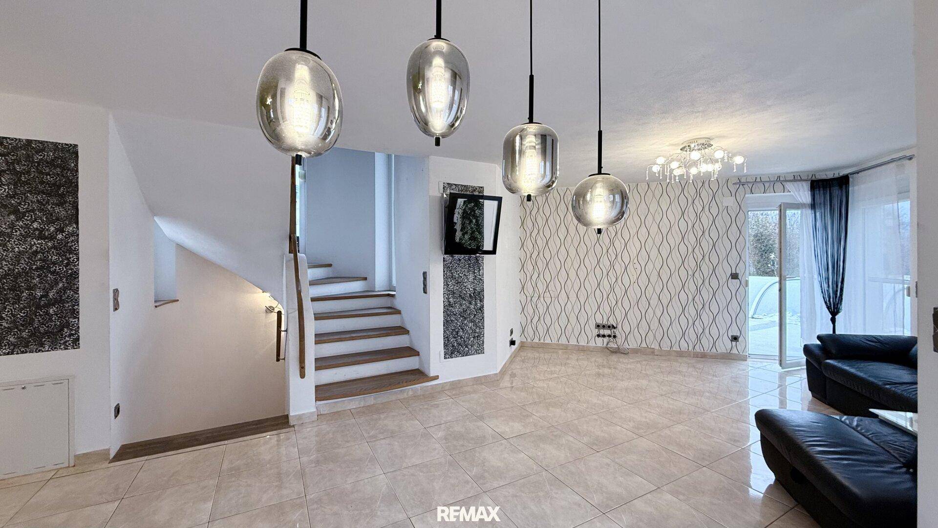 Geräumiges Wohnzimmer mit Fliesenboden, moderner Beleuchtung und Treppe zum Obergeschoss.