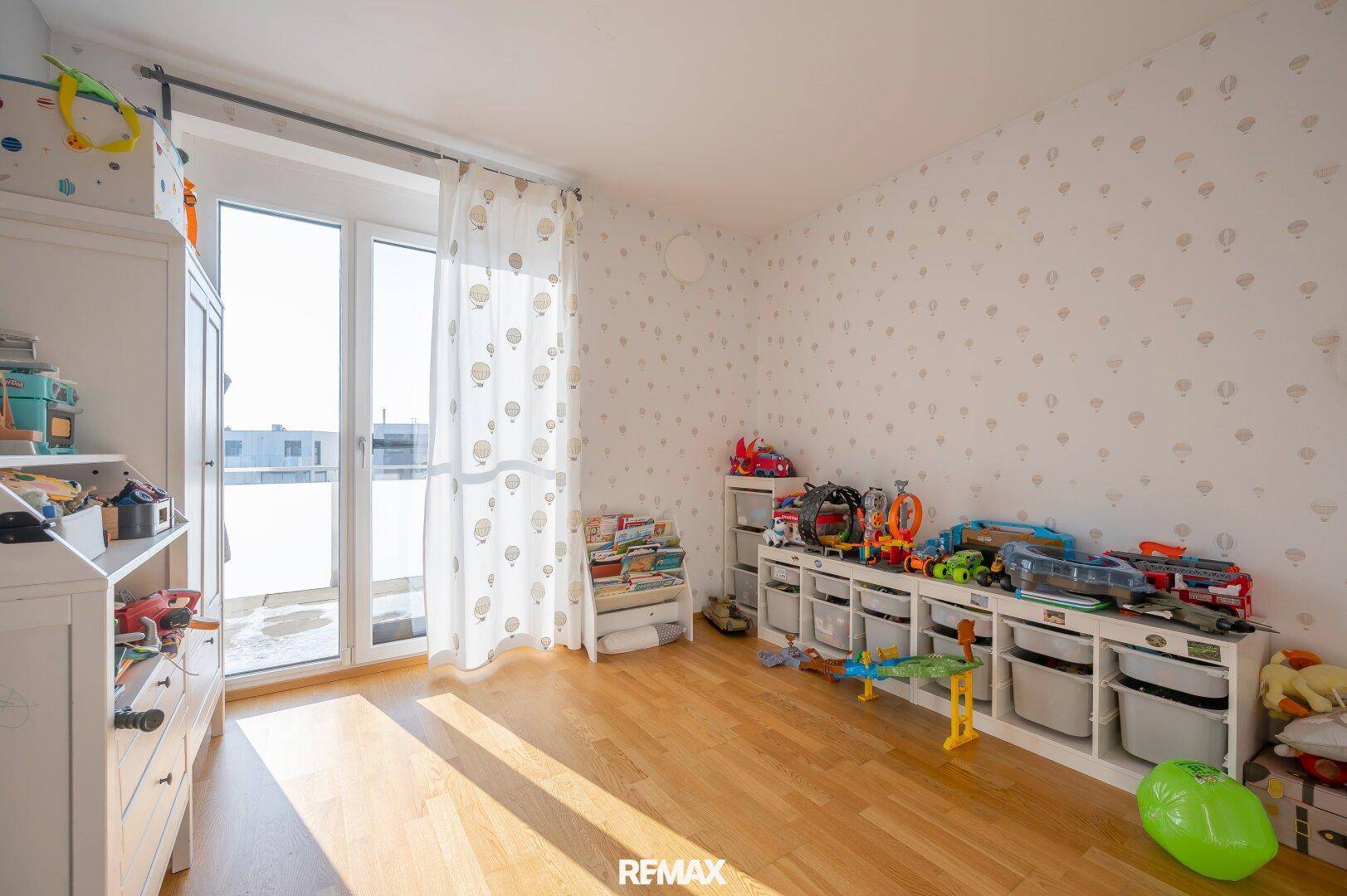 Helles Kinderzimmer mit Holzboden, Spielzeug und Zugang zu einem kleinen Balkon.