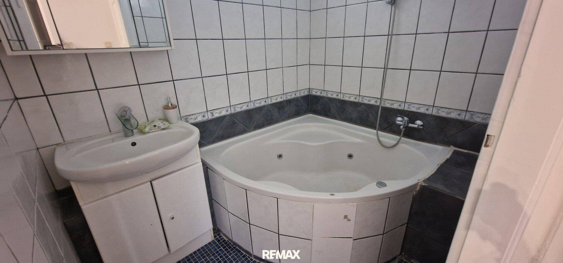Badezimmer mit Eckbadewanne und Duschfunktion, gefliest in Weiß und Dunkelgrau.