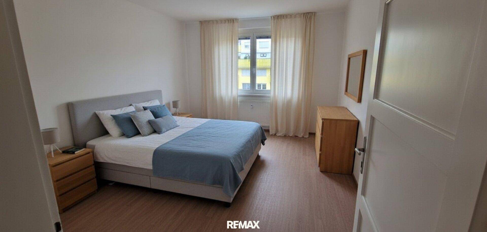 Schlafzimmer mit Doppelbett, Nachttischen und Holzboden, hell durch große Fenster.