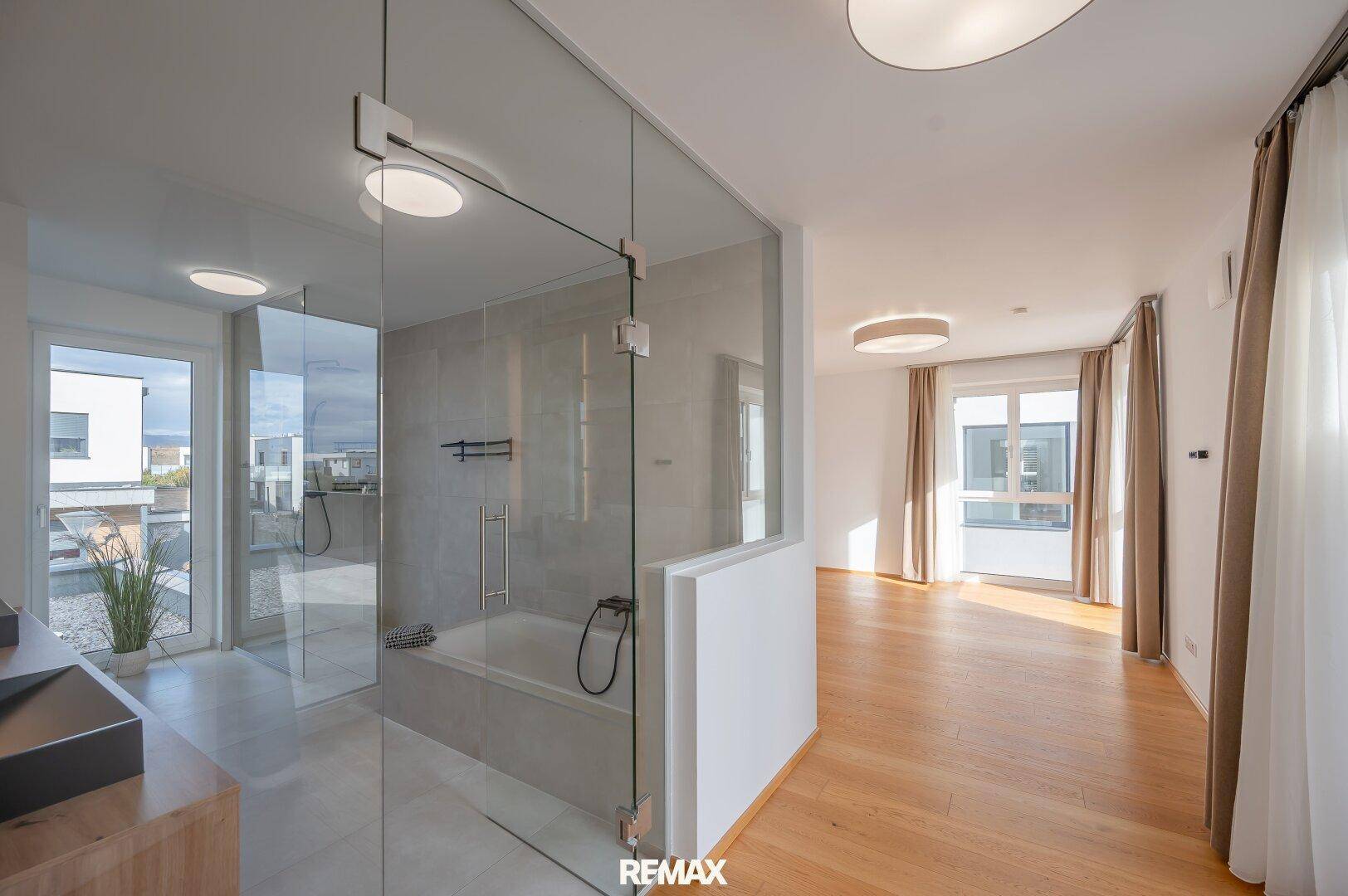 Offenes Badezimmer mit Glasdusche, Badewanne und Holzboden, verbunden mit dem Schlafbereich.