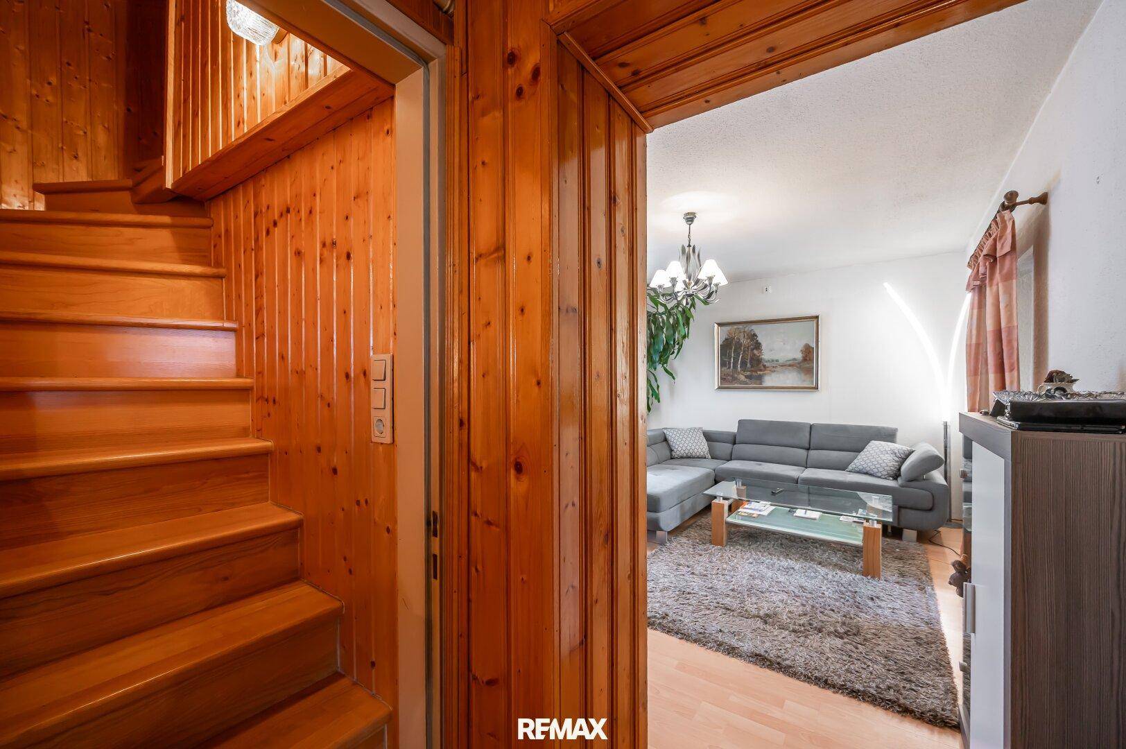 Holztreppe führt zu den oberen Etagen, mit einem Blick in das angrenzende Wohnzimmer mit grauem Sofa.