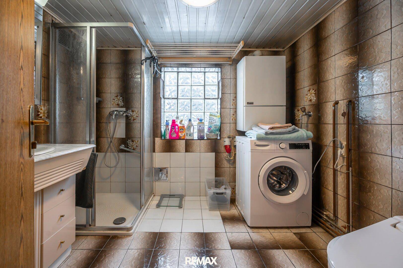 Funktionales Badezimmer mit braunen Fliesen, Duschkabine, Waschbecken und Waschmaschine, gut beleuchtet.