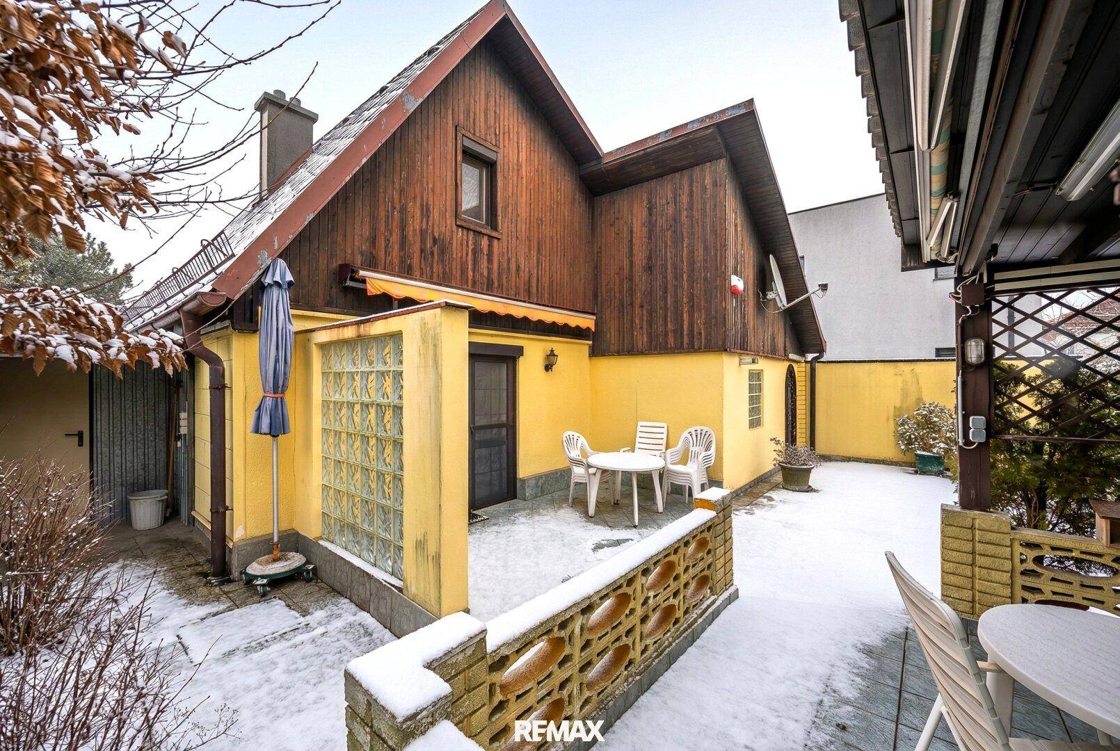 Verschneite Terrasse hinter dem Haus mit Gartenmöbeln und einem Sonnenschirm, bereit für den Frühling.