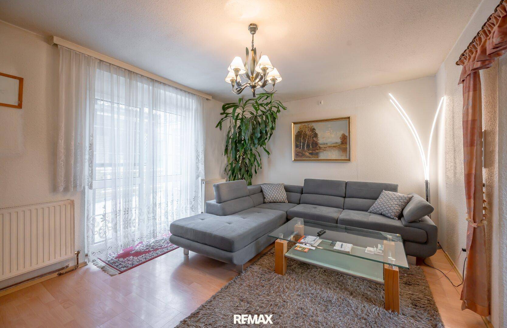 Helles Wohnzimmer mit modernem grauem Ecksofa, Glastisch und großen Fenstern für viel Tageslicht.