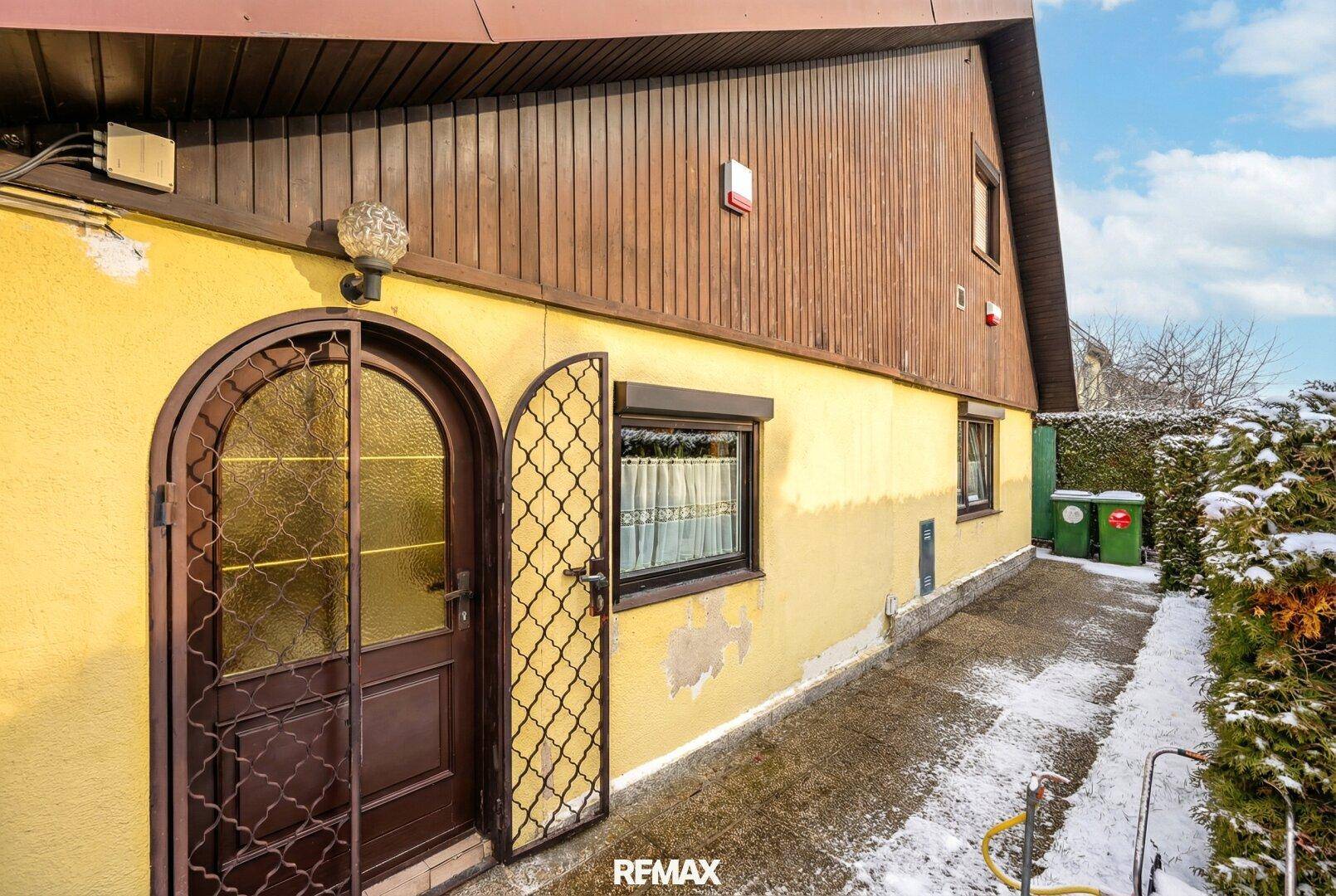 Seitenansicht des Hauses mit gewölbter Eingangstür, Fenster mit Rollläden und verschneitem Zugangsweg.