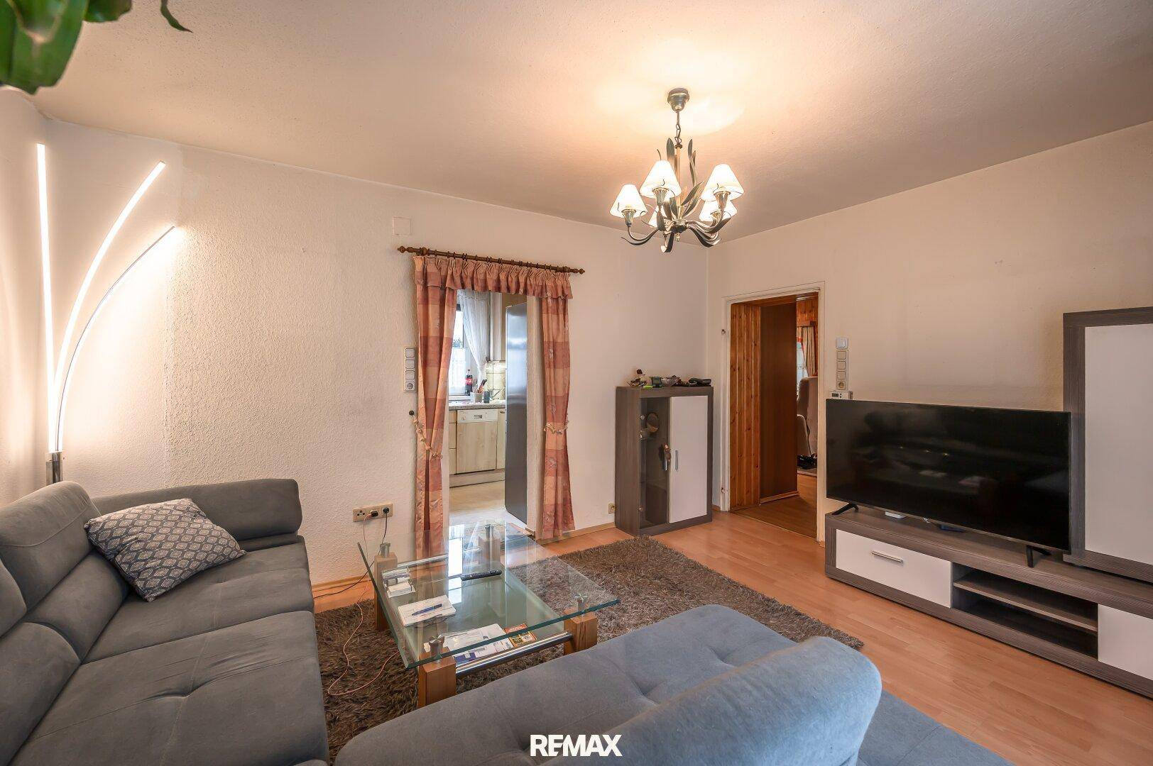 Gemütliches Wohnzimmer mit grauem Sofa, TV-Möbel und Durchgang zur angrenzenden Küche, hell beleuchtet.
