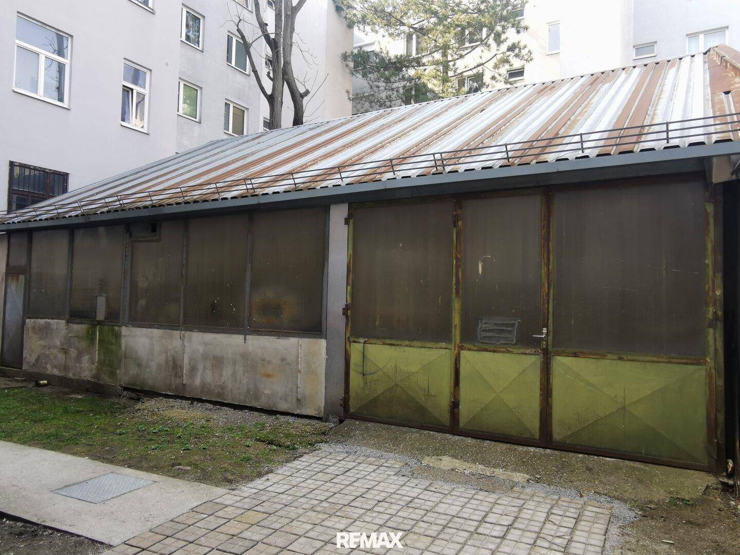 Außenansicht eines langen Gebäudes mit Wellblechdach und großen, undurchsichtigen Fenstern, möglicherweise eine Werkstatt oder Garage.