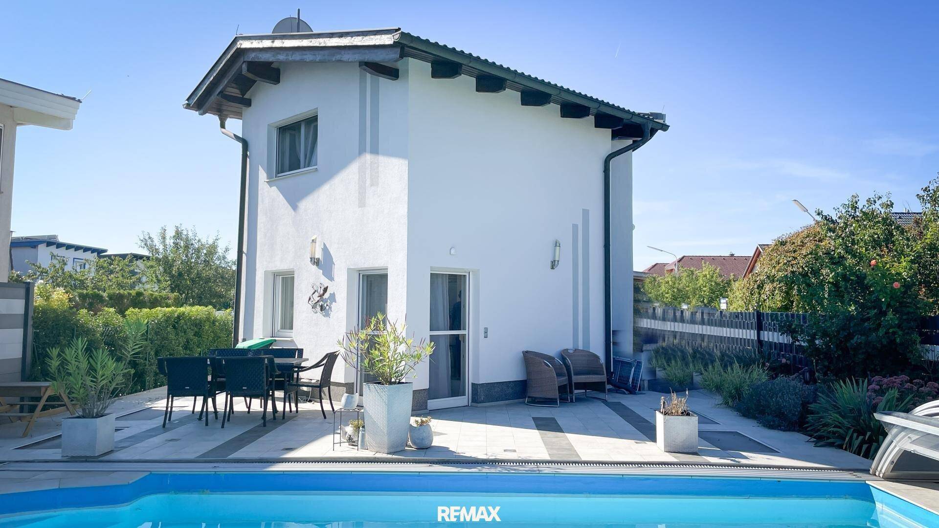 Ansicht des Einfamilienhauses von der Poolseite mit gepflasterter Terrasse und Gartenmöbeln.