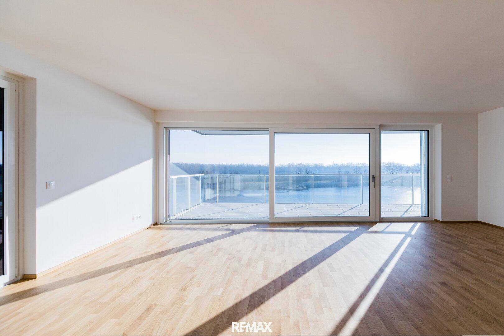 Geräumiges, leeres Wohnzimmer mit Holzboden und großen Fenstern, die viel Tageslicht und Seeblick bieten.