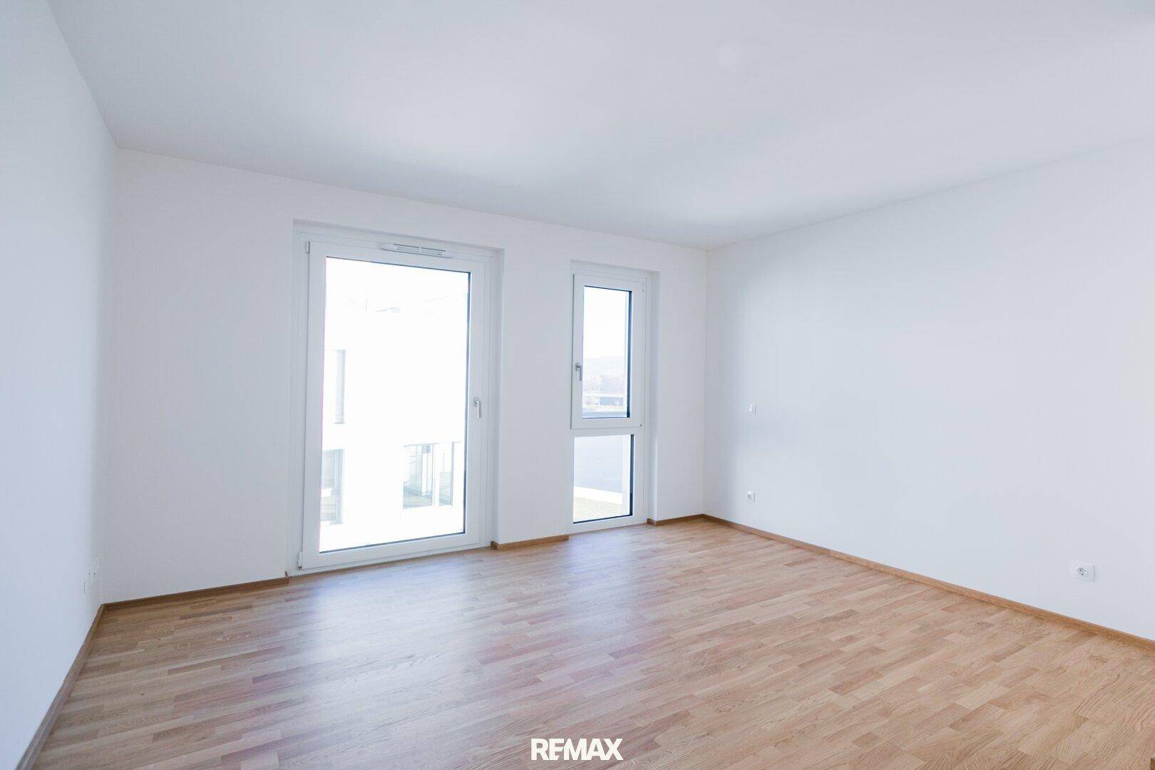 Helles, leeres Zimmer mit Holzboden und Fenstern, die viel Tageslicht hereinlassen und einen Ausblick bieten.