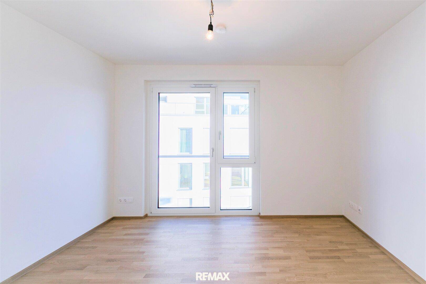 Geräumiges, leeres Zimmer mit Holzboden und großen Fenstern, die den Raum mit natürlichem Licht füllen.