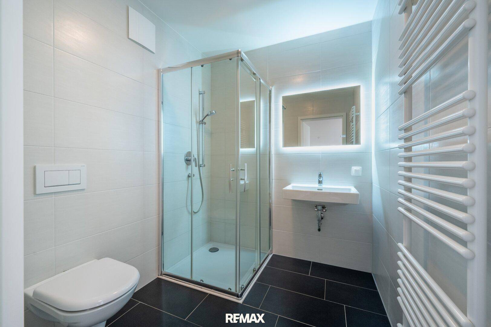 Modernes Badezimmer mit Duschkabine, Wand-WC, beleuchtetem Spiegel und beheiztem Handtuchhalter.
