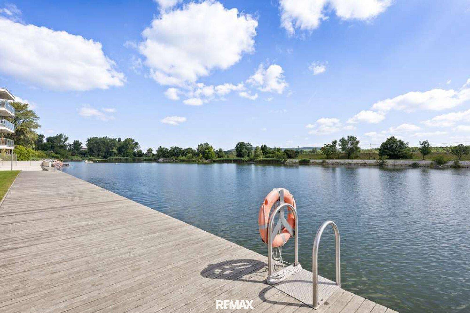 Holzsteg am Flussufer mit Rettungsring, umgeben von grüner Natur und blauem Himmel.