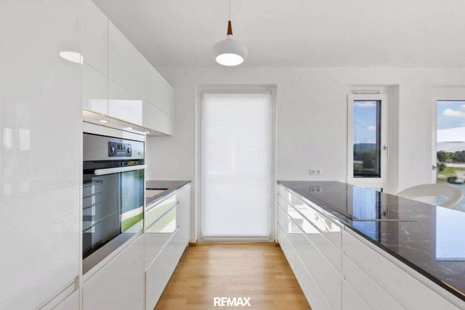 Moderne Küche mit weißen Hochglanzfronten, dunkler Arbeitsplatte und integrierten Geräten, hell durch Fenster.