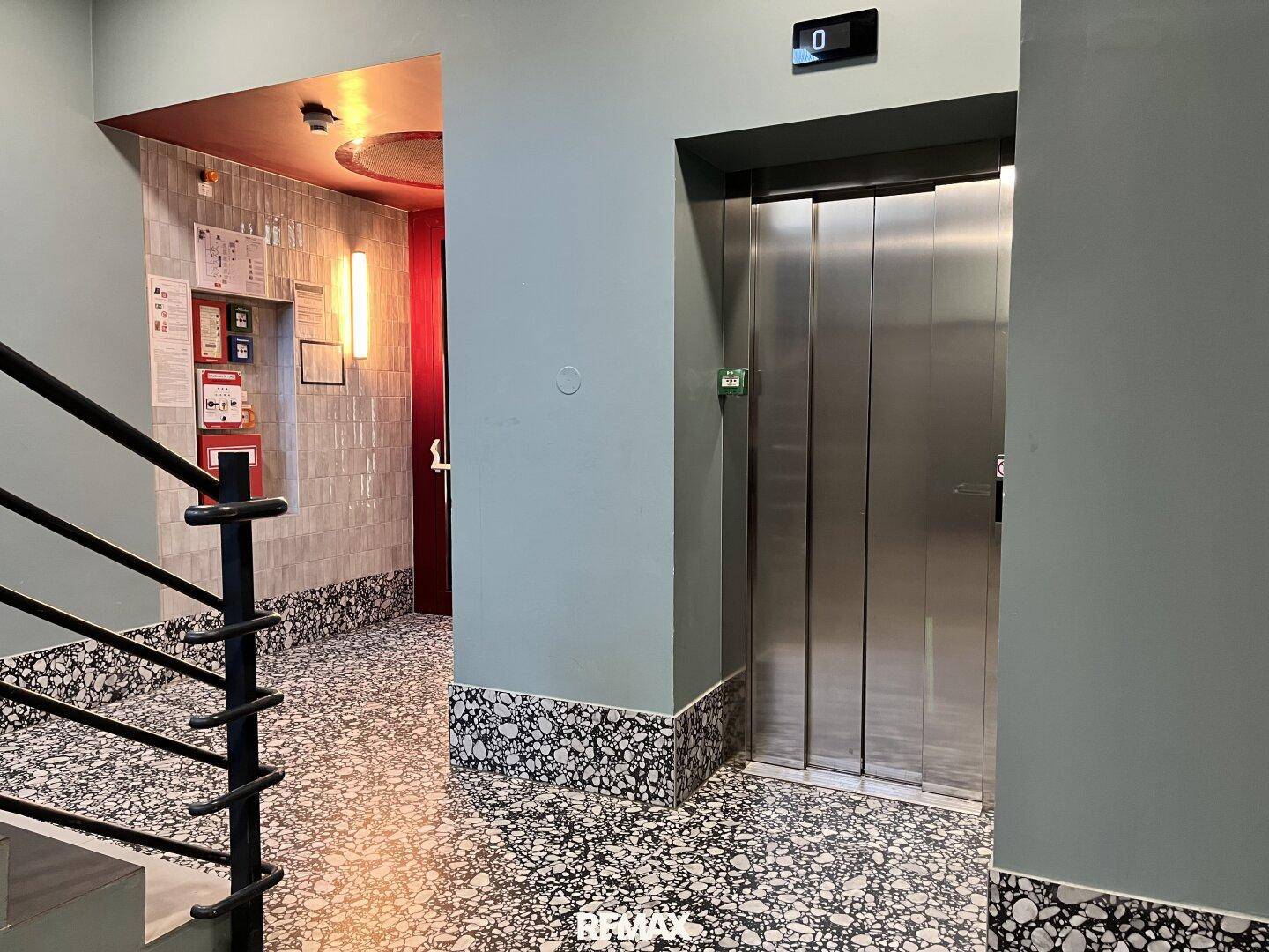 Moderner Eingangsbereich mit Aufzug, Treppe und gemustertem Boden, der Zugang zu den Etagen bietet.