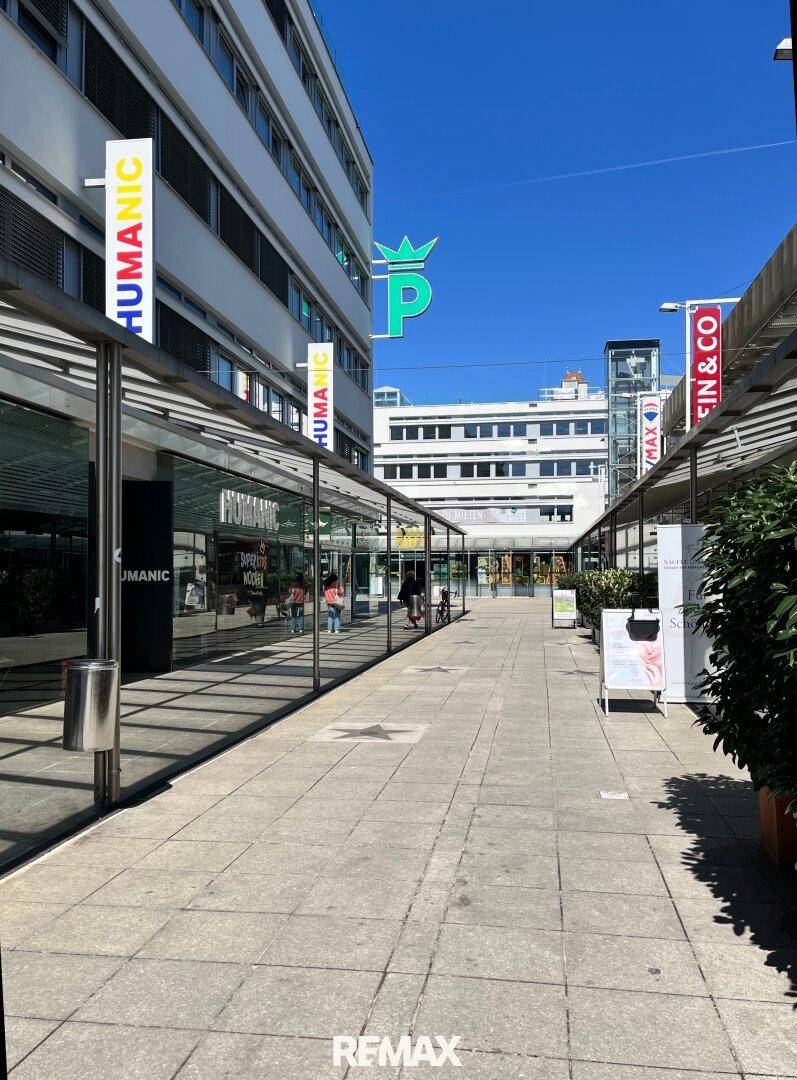 Fußgängerzone zwischen modernen Gebäuden mit Glasfassaden und Geschäften unter blauem Himmel.