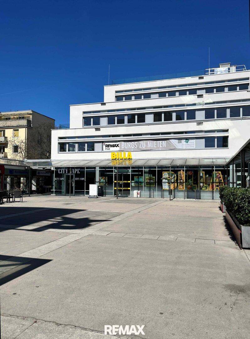 Modernes Geschäftsgebäude mit Supermarkt und weiteren Läden unter strahlend blauem Himmel.