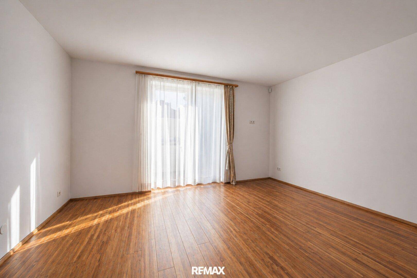 Helles, leeres Zimmer mit Holzboden und großem Fenster, das viel natürliches Licht hereinlässt.