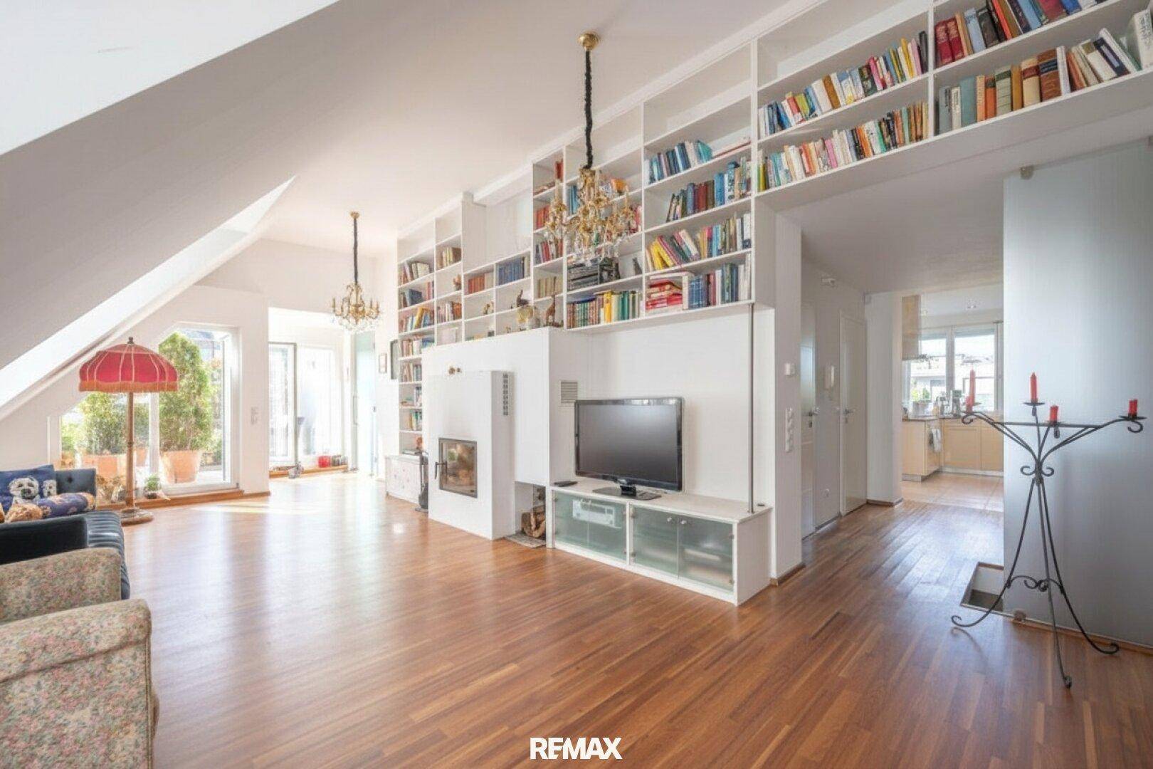 Großzügiges Wohnzimmer mit Holzboden, Einbauregalen, Kamin und eleganten Kronleuchtern unter Dachschräge.