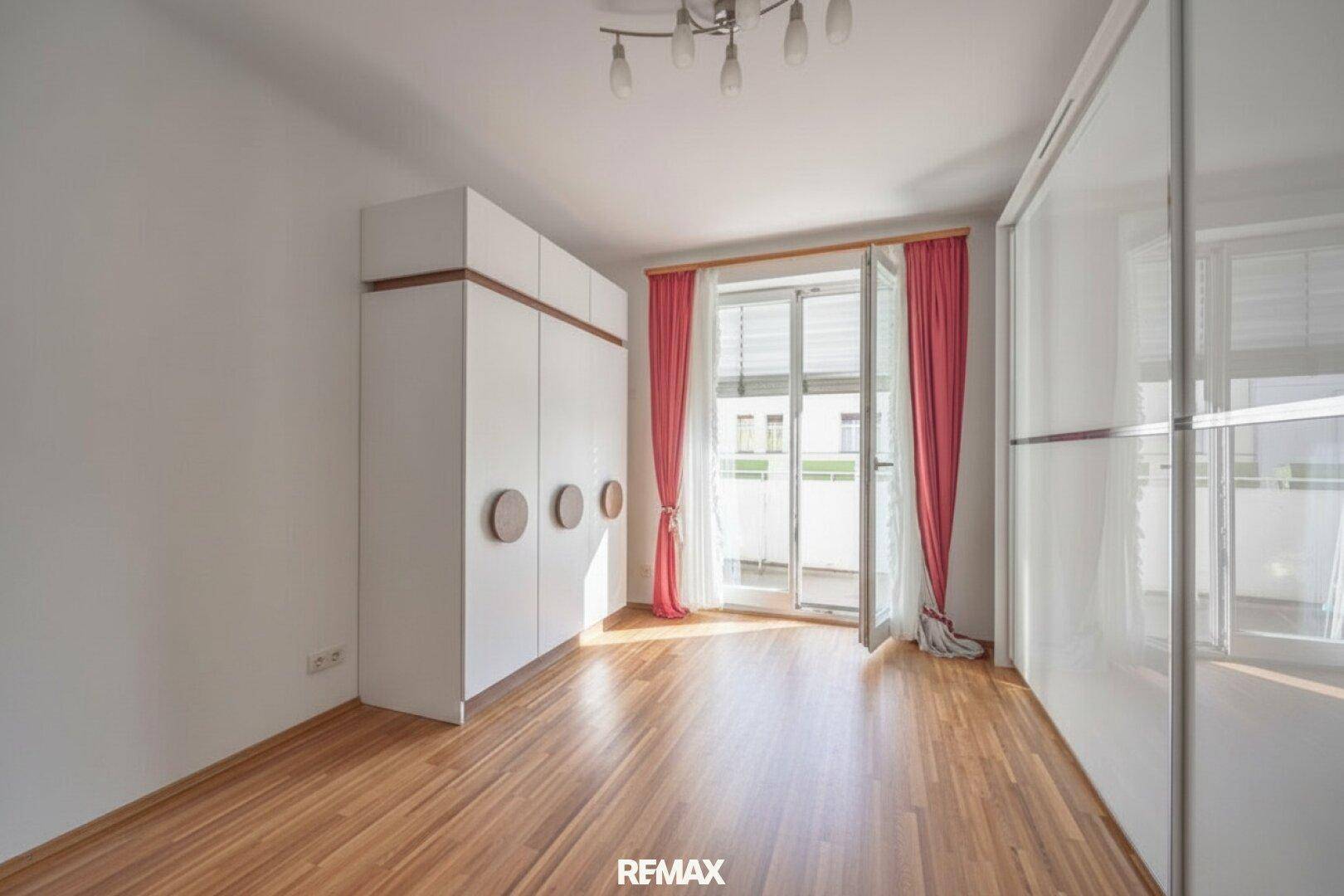 Schlafzimmer mit Holzboden, weißem Kleiderschrank und Zugang zu einem Balkon durch eine Fenstertür.