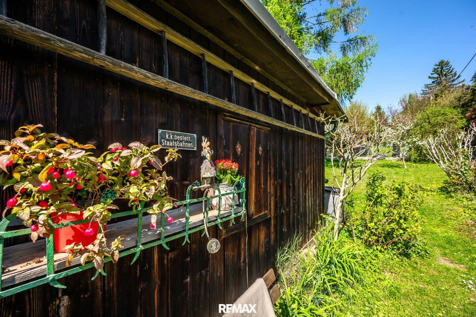 Rustikales Holzgebäude mit Blumenkästen und einem historischen Schild, eingebettet in die grüne Gartenlandschaft.