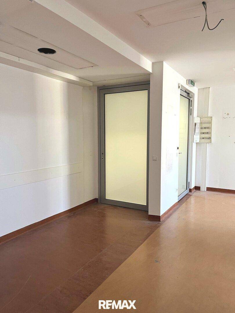 Heller Eingangsbereich mit dunklem Bodenbelag und einer modernen Glastür, daneben ein Sicherungskasten.