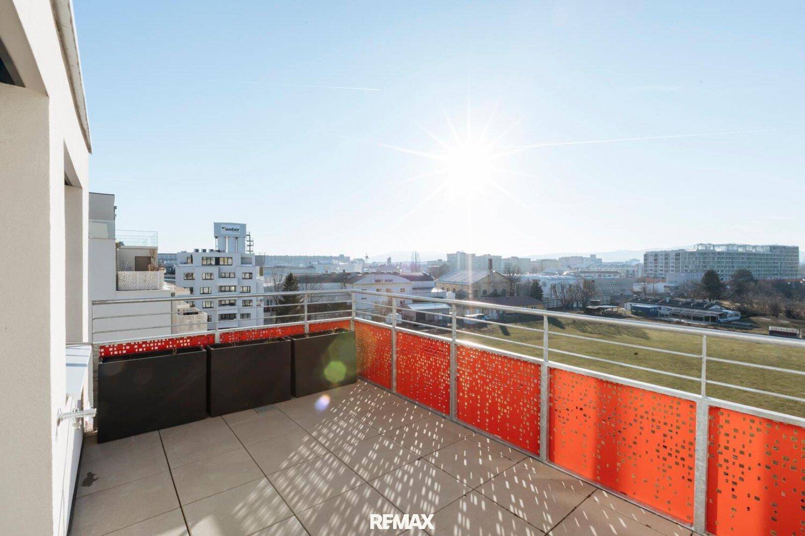 Sonnige Terrasse mit urbanem Ausblick und modernen orangefarbenen Brüstungselementen.