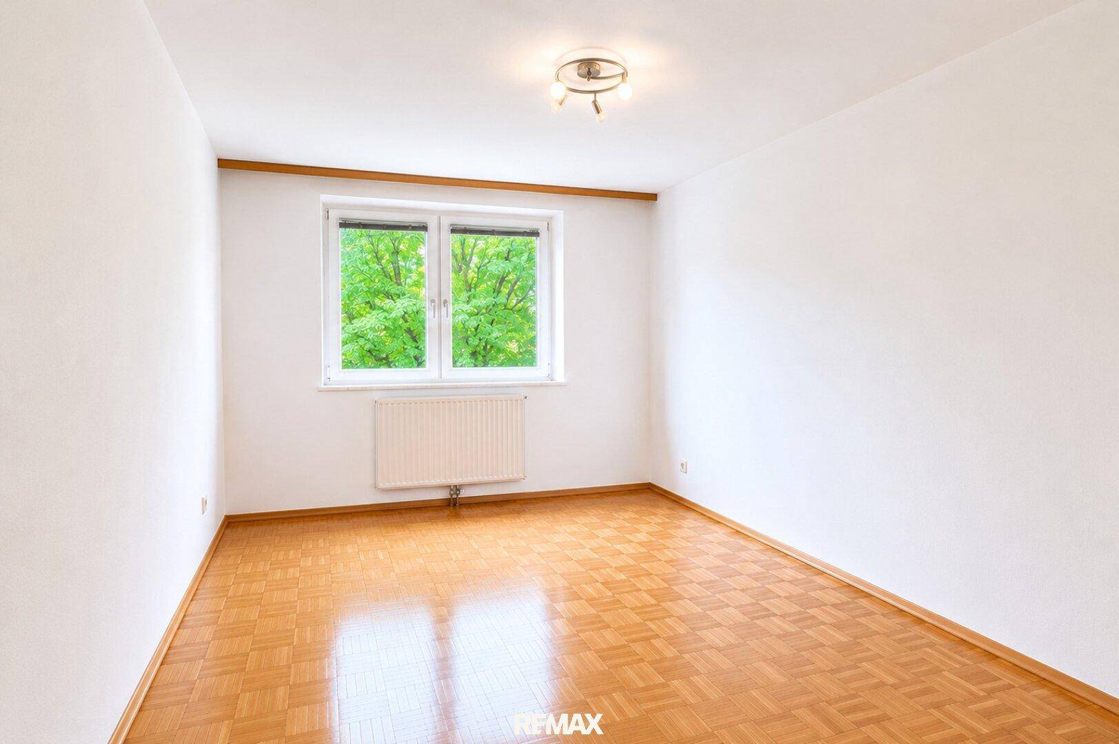 Freundliches Zimmer mit hellem Parkettboden, weißen Wänden und Ausblick ins Grüne.