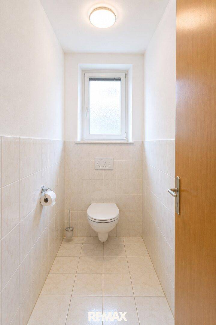 Separates WC mit wandhängender Toilette, hellen Fliesen und einem Fenster für Tageslicht.